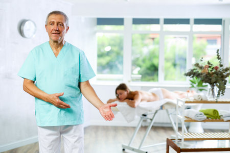 Positive old masseur posing against background of therapy roomの写真素材