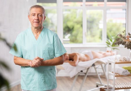 Positive old masseur posing against background of therapy roomの写真素材