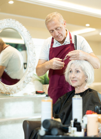 Experienced senior man master makes performs haircut for mature woman with scissorsの写真素材