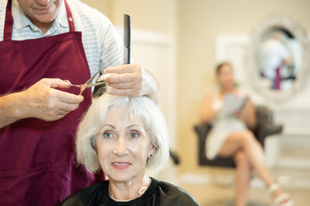 Elderly silver-haired lady getting haircut by male hairdresser using scissorsの写真素材