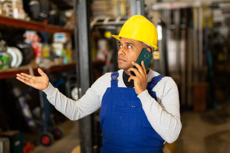 Foreman in protective helmet and overalls talking on cell phone in warehouse of hardware storeの写真素材