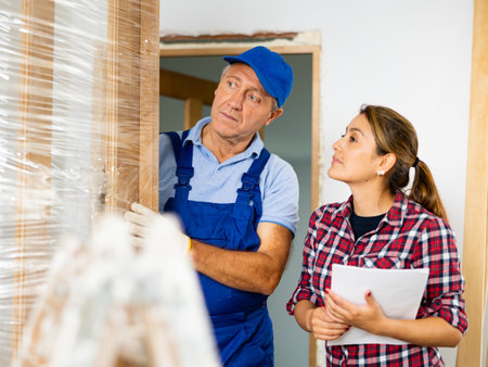 Female in casual clothes, talking with the middle-aged craftsman in uniform while he is holding a wooden door in the houseの写真素材