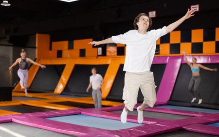 Smiling young male in white t-shirt and beige shorts having great time while jumping on colorful trampoline in game clubの写真素材