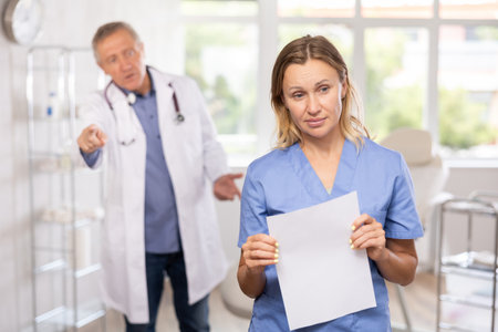 Frustrated female doctor with papers standing in medical office with angry senior colleagueの写真素材