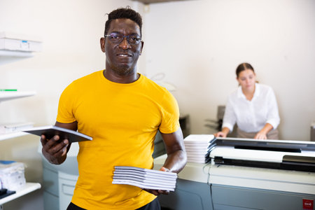 Happy middle-aged African American male typographer showing new notepads in the middle of the copy...