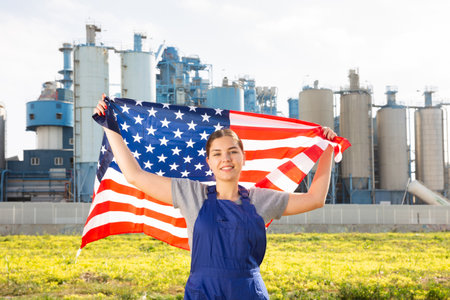 Happy young woman worker with flag of the USA against background of factoryの写真素材