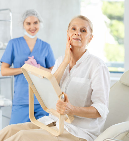 Happy elderly woman examining face after procedures in cosmetology clinicの写真素材