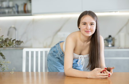 Girl stands in kitchen and holds smartphone in hands. woman reads news and likes photos of friends on social networksの写真素材