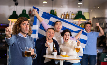Group of emotive friends supporters cheering for favorite team with flag of Greece while watching match on TV in pubの写真素材