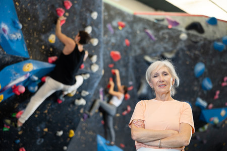 Elderly woman in sportswear smiling at camera while standing against artificial training climbing rock wall in adventure parkの写真素材