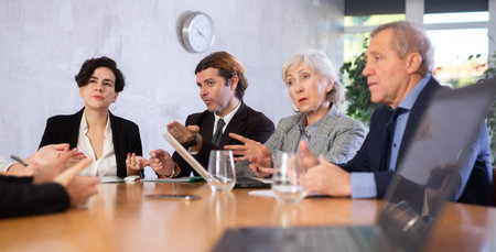 Office coworkers of different ages discussing topic of day while sitting at table with laptop and...