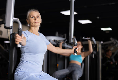 Elderly female training trains muscles of arms and press on simulator in gymの写真素材