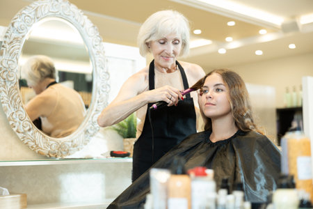 Female hairdresser makes curls the hair of young woman using curling ironの写真素材