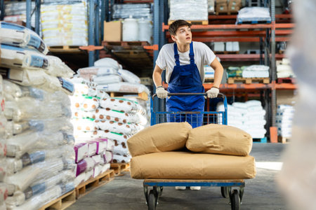 In warehouse of store, stevedore loader young man pushes and carries large cart for bulky cargo
