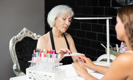 Friendly elderly woman working in beauty salon as manicure master shaping the nails of young female client with nail fileの写真素材