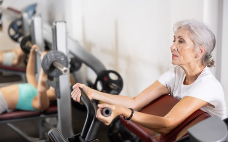 Slender senior woman work out muscles of arms and shoulders on simulator in gym of fitness club.の写真素材