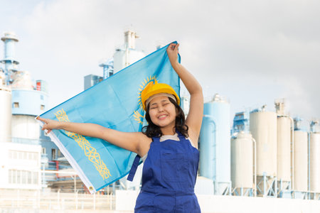 National flag of Kazakhstan in the hands of girl in overalls against background of modern metallurgical plantの写真素材