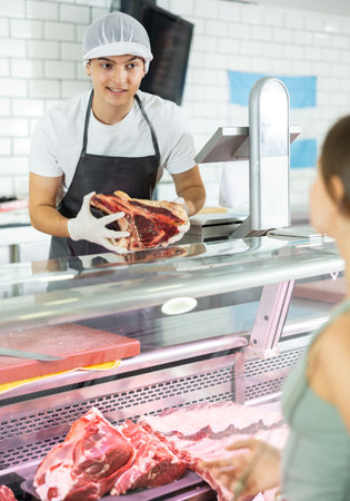 Positive young salesman demonstrating piece of meat to purchaser in butcher shopの写真素材