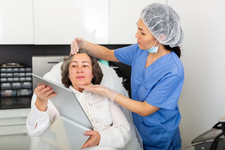 Young female cosmetologist gave an elderly woman client to admire in the mirror, examining her before the procedureの写真素材