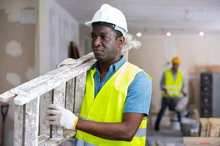 African american man builder in yellow vest carrying ladder while working in room