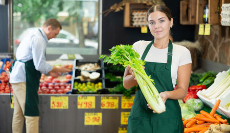 Young female seller in apron displaying assortment of celery at supermarketの写真素材