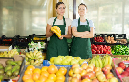 Friendly woman owner of fruit-vegetable shop standing in trading floor with girl assistantの写真素材