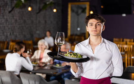Young waiter guy is standing with order tray in spacious guest room in restaurant.の写真素材