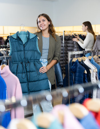 Smiling female woman keeping turquoise winter waistcoat on a rack while shopping in the showroomの写真素材