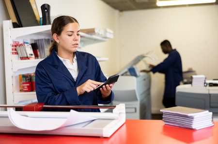 Attentive young woman in a blue uniform calculating the cost of the work done in the printing houseの写真素材