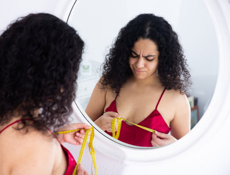 Dissatisfied woman measuring bust size near mirrorの写真素材