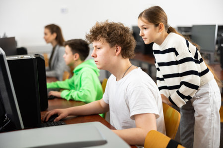 Teen schoolgirl helping focused classmate sitting at computer in classの写真素材