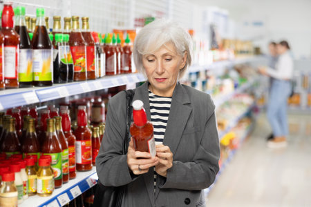 Senior woman customer is thinking about buying spicy and unusual red chilli tsuyu sauce.の写真素材