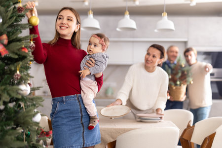 Young woman with a baby in her arms decorates a Christmas tree with toys while the rest of the family prepares festive dinnerの写真素材