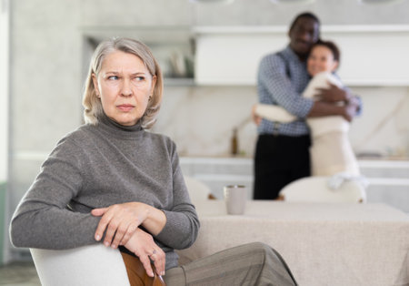 Sad old woman sitting at the kitchen table with her back to couple posing happily in the kitchenの写真素材