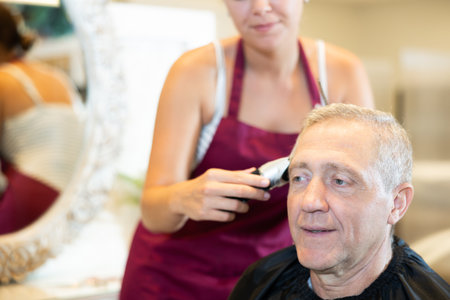Cropped young woman hairdresser performs haircut for elderly man using hair clipper.の写真素材