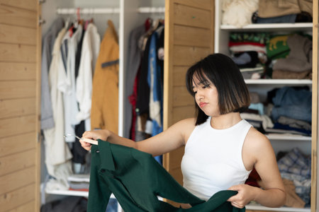 Sad young Asian girl trying to choose a dress standing by wardrobe