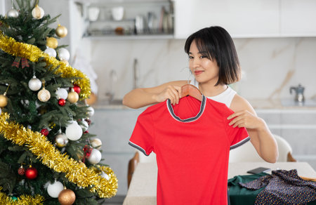Happy young Asian girl trying to choose a dress standing next to Xmas-treeの写真素材