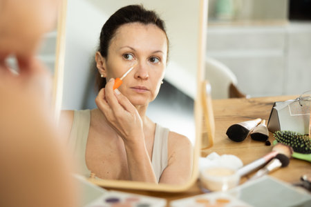 Middle-aged woman applying concealer on her facial skin sitting in front of the mirrorの写真素材