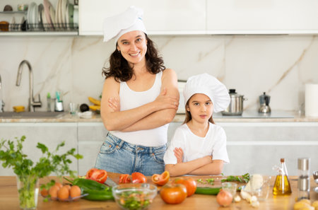 Mother and girl in white cooking hats are standing in kitchen near tableの写真素材