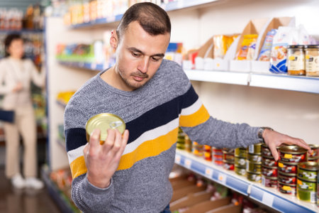 Man chooses canned fish in grocery store. Man shopper reading expiration date on product labelの写真素材
