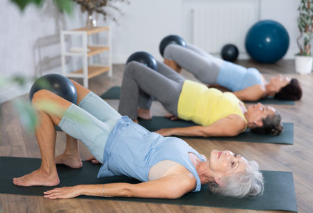 Old woman practicing bridge pose with softball between legs during fitness classesの写真素材