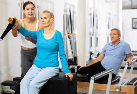 Positive young female instructor controlling movements of mature woman exercising on Pilates reformer equipment in studioの写真素材