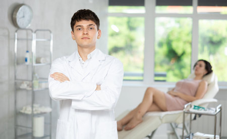 Young man doctor posing in doctors cabinet with patient lying on clinical chairの写真素材