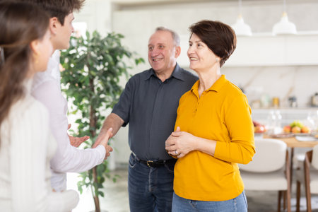 Happy mature man and woman welcoming son and young wife meeting in living room by served dinner tableの写真素材