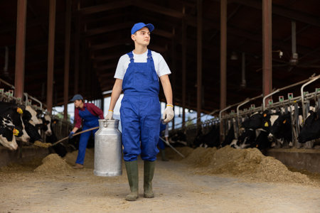 Portrait of young European man dairy farmworker in uniform carrying large metal milk can in cowshedの写真素材
