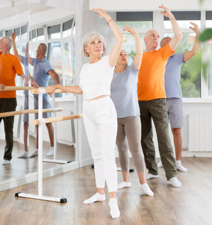 Active mature woman practicing battement tendu holding by ballet barre together with othersの写真素材