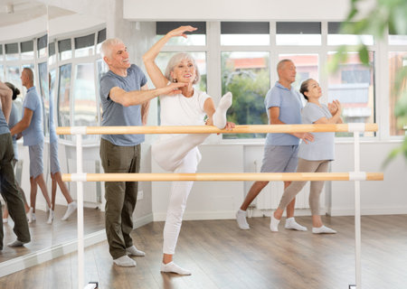 Active mature woman practicing leg stretching holding by ballet barre with instructor