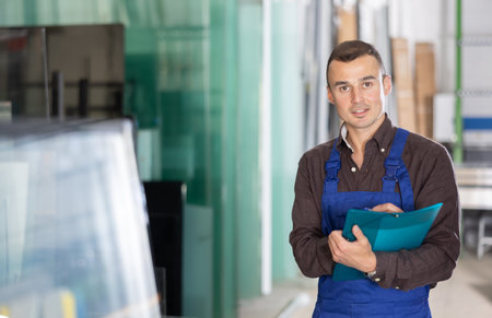 Positive factory worker with clipboard standing in glass workshop