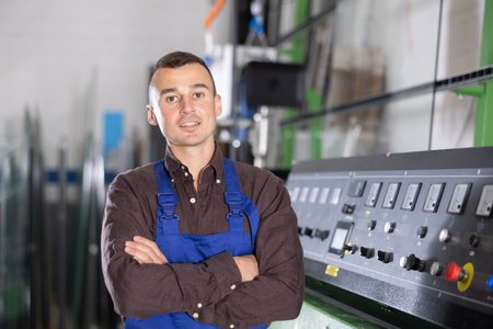 Smiling worker standing near machinery control panel in glass processing workshop