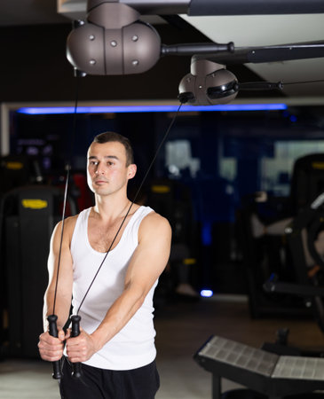 Sportive young guy training his arms on adjustable cable exercise machine in gym